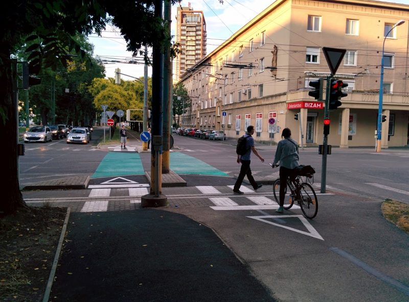 Cyklotrasa R26: Cyklistka čaká na STOP čiare na signál voľno, medzitým chodec prechádza križovatkou.